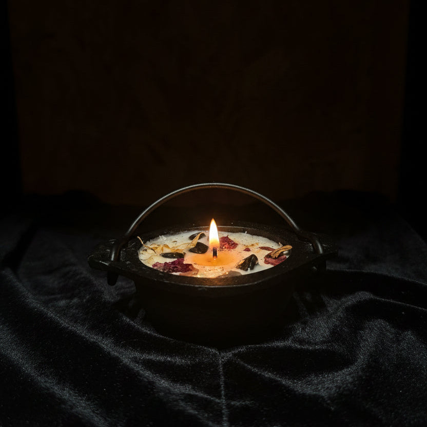 A small black cast-iron cauldron-shaped candle with a handle, filled with cream wax and scattered dried flower petals, resting on a black velvet cloth against a rustic wood panel background.