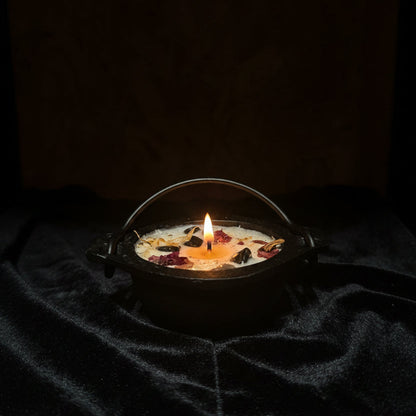 A small black cast-iron cauldron-shaped candle with a handle, filled with cream wax and scattered dried flower petals, resting on a black velvet cloth against a rustic wood panel background.